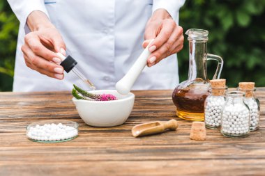 cropped view of woman holding pestle and pipette near mortar with veronica flowers and jug with oil on wooden table  clipart