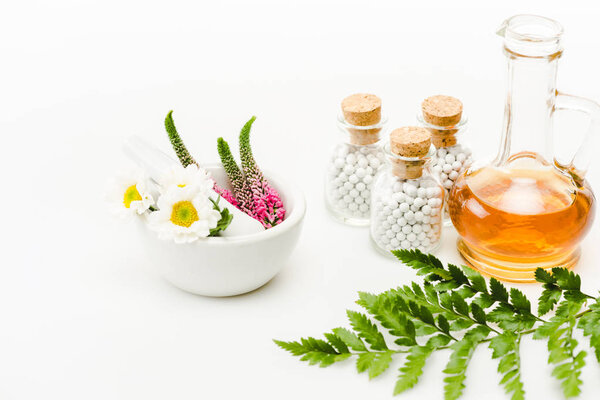 flowers in mortar near pestle and glass bottles with pills, jug with oil and green leaf on white 