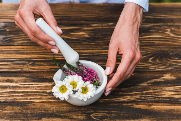 cropped view of woman holding pestle near mortar with flowers on wooden table 