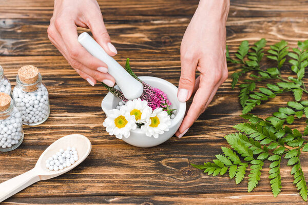 cropped view of woman holding pestle near mortar with flowers and green leaves on wooden table 