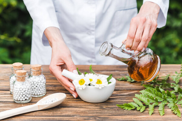 cropped view of woman holding jug with oil near mortar with flowers and green leaves on wooden table 