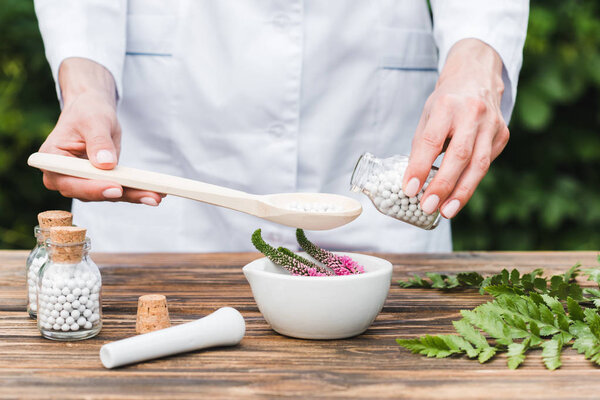 cropped view of woman holding spoon and bottle with pills near mortar with veronica flowers and green leaves on wooden table 
