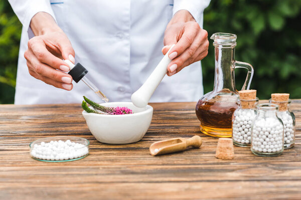 cropped view of woman holding pestle and pipette near mortar with veronica flowers and jug with oil on wooden table 