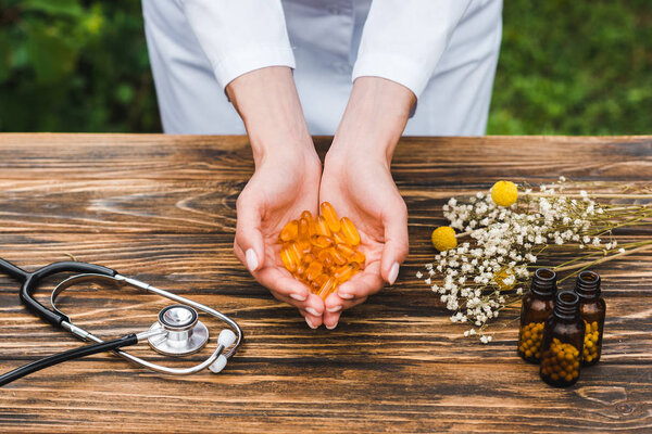 cropped view of woman pills near bottles flowers and stethoscope on wooden table 