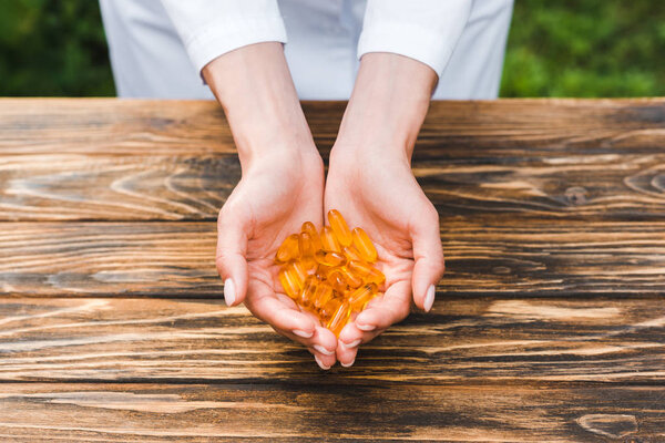 cropped view of woman holding orange pills in hands near wooden table 