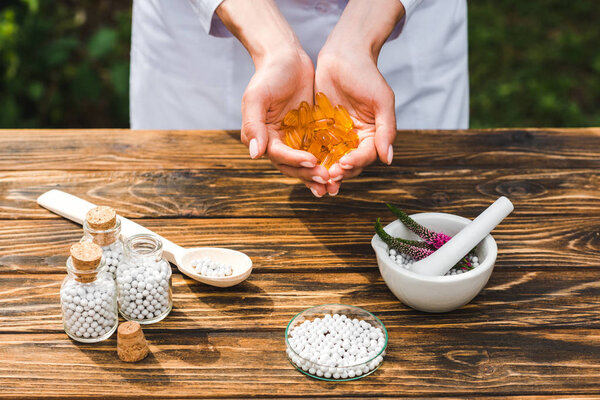 cropped view of woman holding orange pills near bottles and mortar with veronica flowers on wooden table 