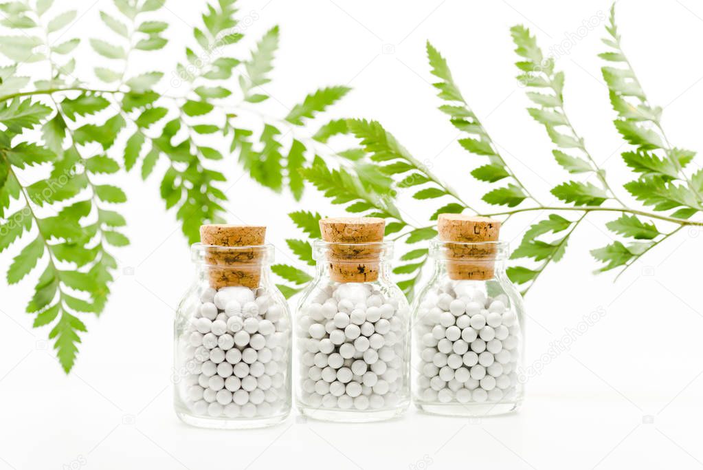 selective focus of small pills in glass bottles with wooden corks near green leaves on white 