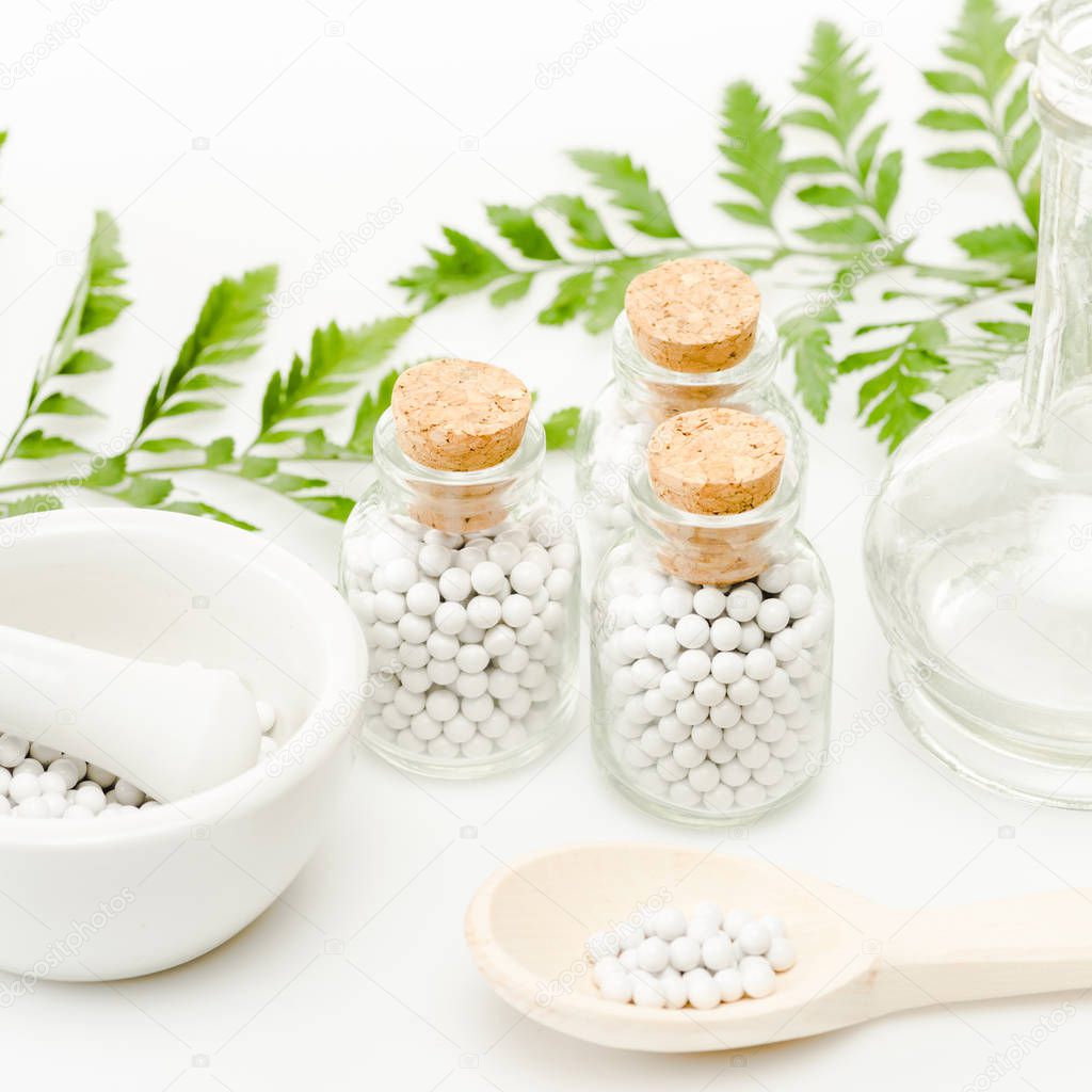 glass bottles with small round pills near mortar and pestle, wooden spoon, jar and green leaves on white 