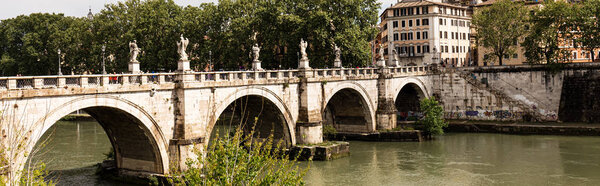ROME, ITALY - JUNE 28, 2019: panoramic shot of river Tiber and people walking on old bridge in sunny day