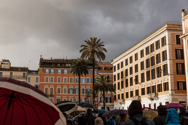 ROME, ITALY - JUNE 28, 2019: people with umbrellas walking past buildings and palm trees under overcast sky