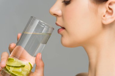 side view of young woman looking at glass of water with sliced lime isolated on grey  clipart
