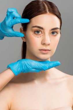 cropped view of plastic surgeon in latex gloves near attractive woman with marks on face isolated on grey  clipart