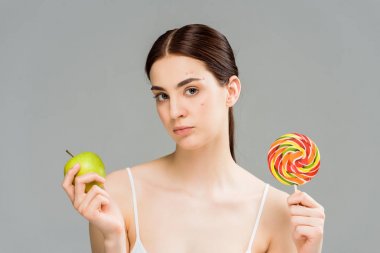 gri izole tatlı lolipop ve yeşil elma tutan yüzünde akne ile esmer kadın 