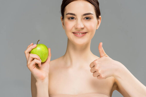 счастливая женщина улыбается, держа зеленое яблоко и показывая большой палец на сером
 