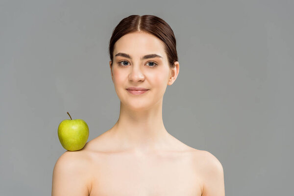 брюнетка обнаженная женщина с зеленым яблоком на плече улыбается изолированно на сером
 