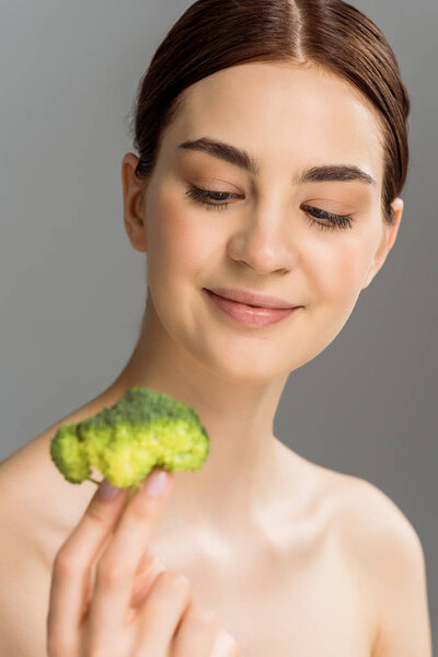 selective focus of happy naked woman smiling while holding green broccoli isolated on grey 