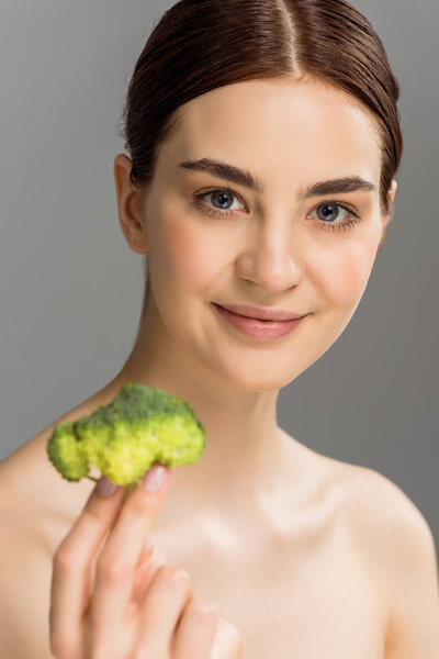 selective focus of cheerful naked woman smiling while holding green broccoli isolated on grey 