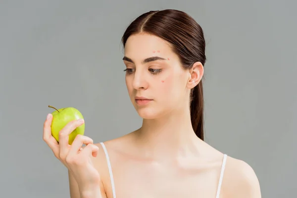 young woman with acne looking at green apple isolated on grey