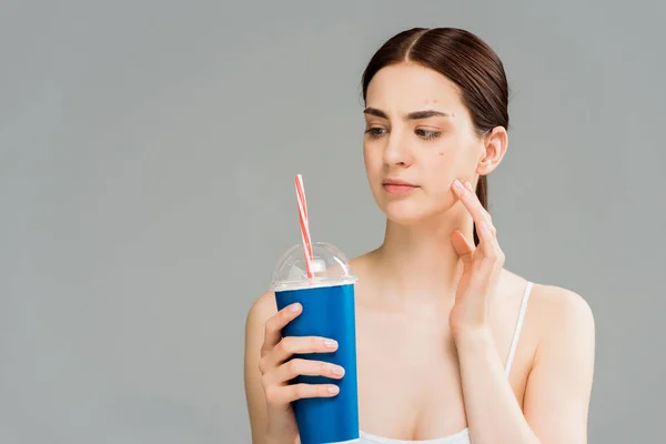 woman with acne on face looking at plastic cup with straw isolated on grey