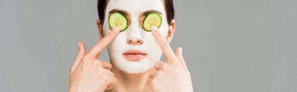 panoramic shot of young woman with clay mask and cucumbers on face isolated on grey 