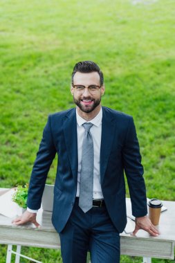 high angle view of smiling businessman standing near table in park, smiling and looking at camera clipart