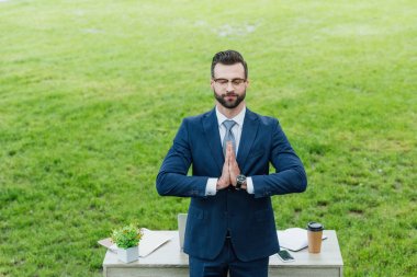 businessman closing eyes and gesturing while standing near white table in park clipart