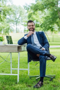 full length view of businessman sitting in office chair near table with laptop and plant, smiling and looking at camera clipart