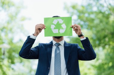 young man covering face with recycle sign card while standing in park clipart