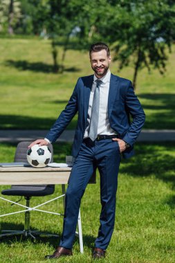 full length view of young businessman putting football on table while standing in park  clipart