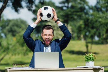 dizüstü bilgisayar, saksı ve gözlük ile masada oturan genç işadamı, gülümseyerek ve baş üzerinde futbol tutarak