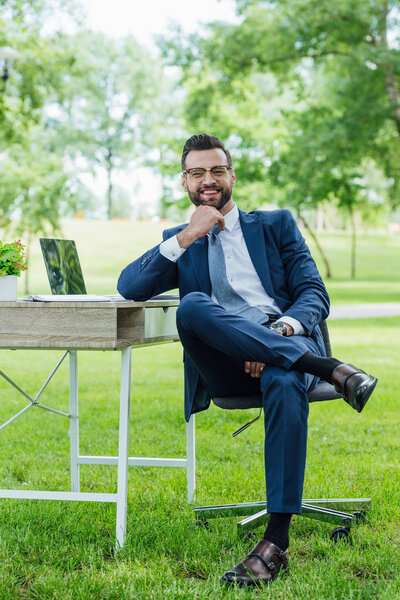 full length view of businessman sitting in office chair near table with laptop and plant, smiling and looking at camera