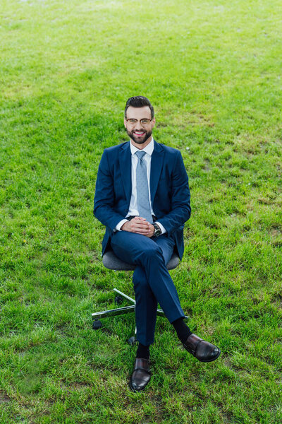 high angle view of happy young businessman sitting in office chair in park and loooking at camera