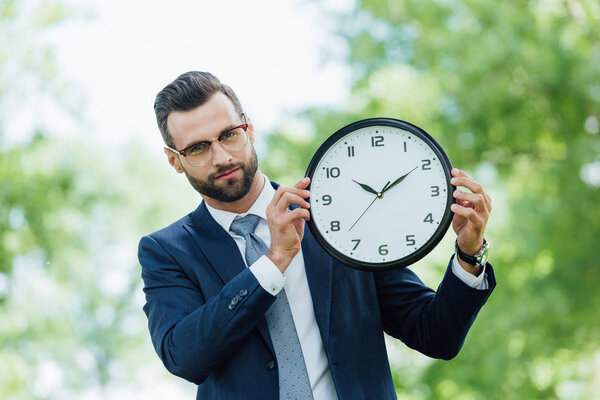 young businessman holding clock and looking at camera while standing in park