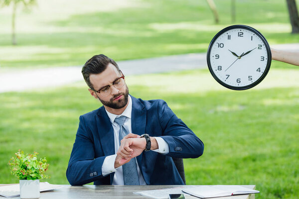 businessman sitting at table with plant in park and looking at watch 