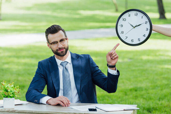 young businessman sitting at table with plant, smartphone and notebooks, looking at camera and pointing with finger at clock