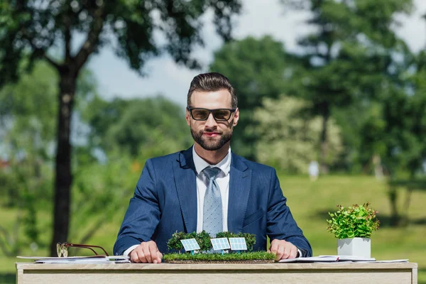 サングラスをかけた若いビジネスマンは 太陽電池のレイアウトと植物と植木鉢とテーブルに座って カメラを見て — ストック写真