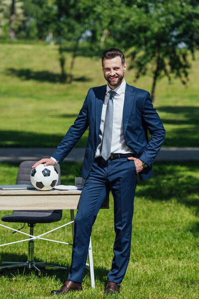 Вид в полный рост, как молодой бизнесмен ставит футбол на стол, стоя в парке
 
