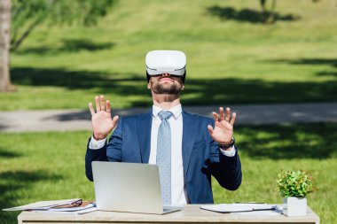 parkta dizüstü bilgisayar, dizüstü bilgisayarlar ve saksı ile masa otururken Vr kulaklık gesturing genç işadamının ön görünümü 
