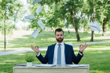 young businessman sitting in park behind table and meditating near flying papers clipart