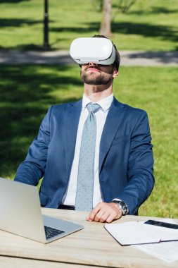 Vr kulaklık lı genç işadamı dizüstü bilgisayar ile masanın arkasında parkta oturuyor