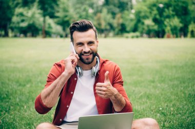 dizüstü bilgisayar ile çim üzerinde oturan genç adam, akıllı telefon üzerinde konuşurken ve başparmak gösteren