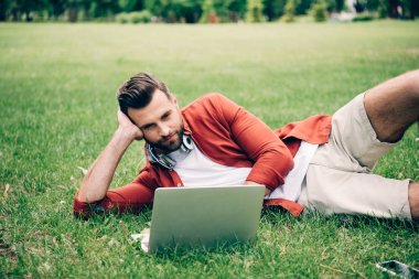parkta çimüzerinde yatan ve dizüstü bilgisayar kullanan genç adam