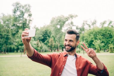 parkta duran yakışıklı adam, gülümseyerek, selfie çekerek ve zafer işareti gösteren