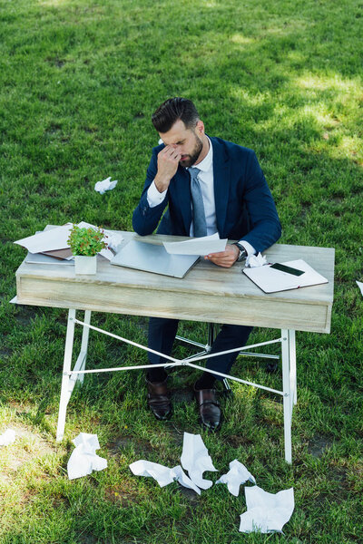 tired businessman sitting in park behind table with laptop, smartphone, notebooks and flowerpot with many crumpled sheets of paper on grass