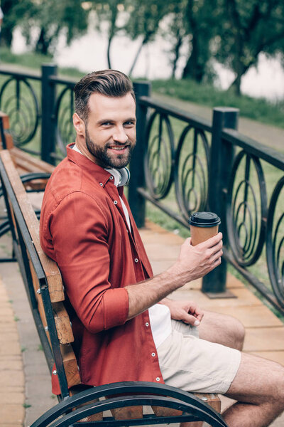 young man in red shirt sitting on bench, holding coffee to go, smiling and looking at camera