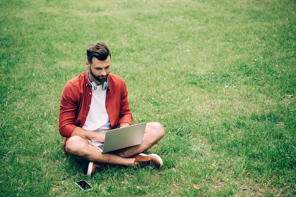 Człowiek Siedzi Trawie Parku Pobliżu Smartphone Pomocą Laptopa — Zdjęcie stockowe