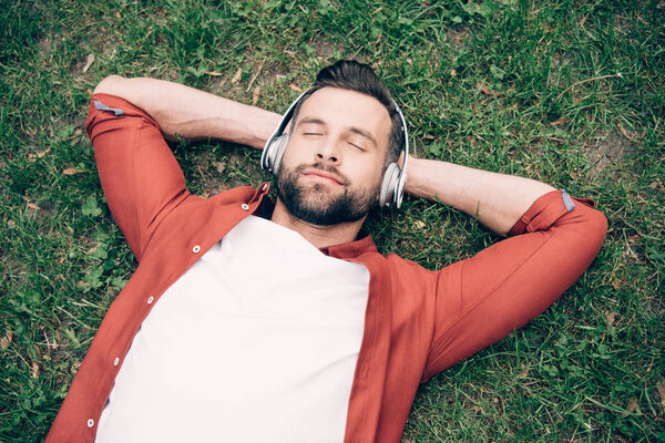 young man with closed eyes lying on grass with hands behind head and listening to music