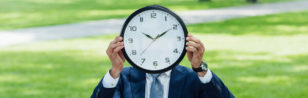 panoramic shot of businessman covering face with clock while standing in park