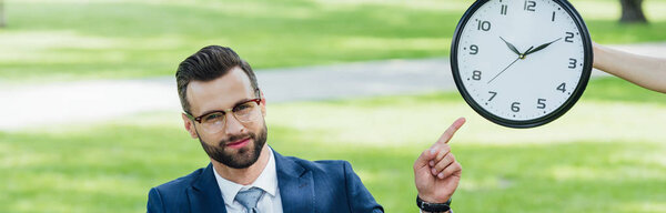 panoramic shot of businessman pointing with finger at clock while sitting in park