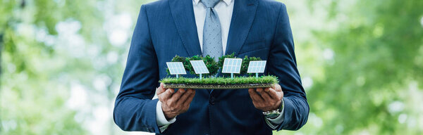 панорамный снимок бизнесмена, держащего в руках макет солнечных батарей, стоя в парке
